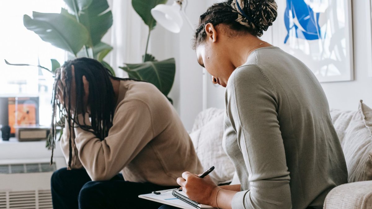 Professional female counselor taking notes in notepad while listening to depressed African American male client touching head in dismay in modern psychology center