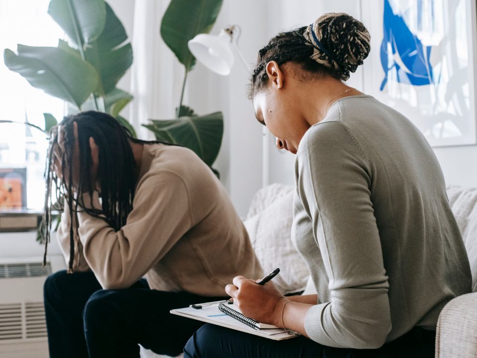 Professional female counselor taking notes in notepad while listening to depressed African American male client touching head in dismay in modern psychology center